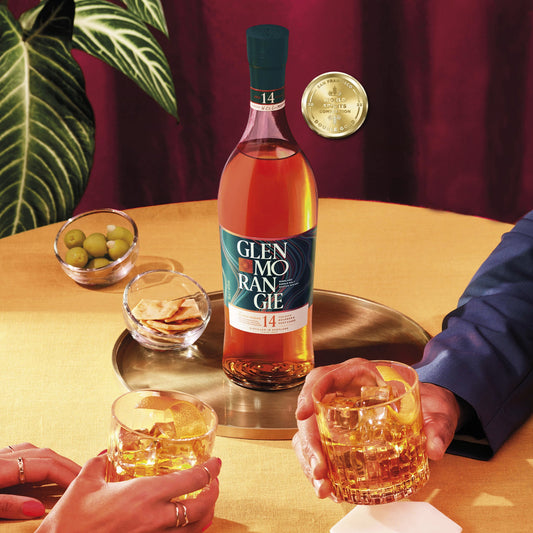 Glenmorangie whisky bottle with two glasses on a table, surrounded by decorative elements.